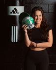A woman in a black athletic outfit stands in front of a black brick wall, holding a green and silver boxing glove. A black Adidas punching bag hangs on the wall behind her.