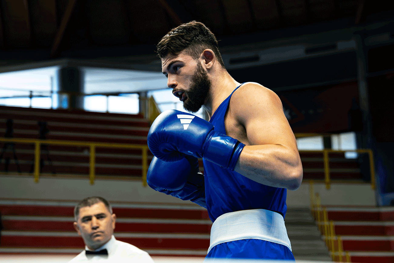 Man in boxing ring with blue adidas Combat Sports boxing gloves. Referee in background. 