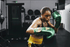 A female boxer in a fighting stance wearing metallic green and gold Adidas boxing gloves, a black sports bra, and gold boxing shorts. She gazes intensely at the camera with both gloves raised in a guard position.