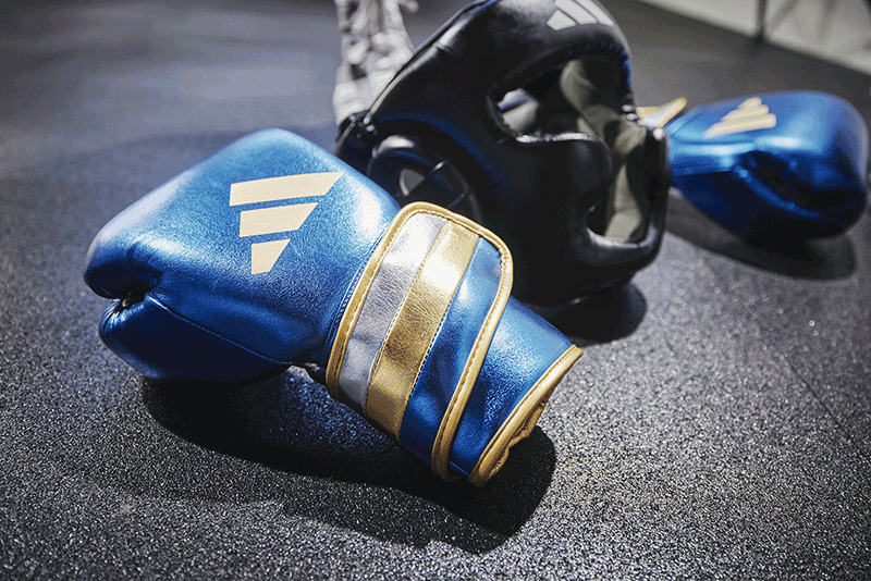 A pair of metallic blue and gold Adidas boxing gloves resting on a gym floor, with a black Adidas boxing headguard and a second blue glove visible in the background.