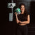 A woman in a black athletic outfit stands in front of a black brick wall, holding a green and silver boxing glove. A black Adidas punching bag hangs on the wall behind her.
