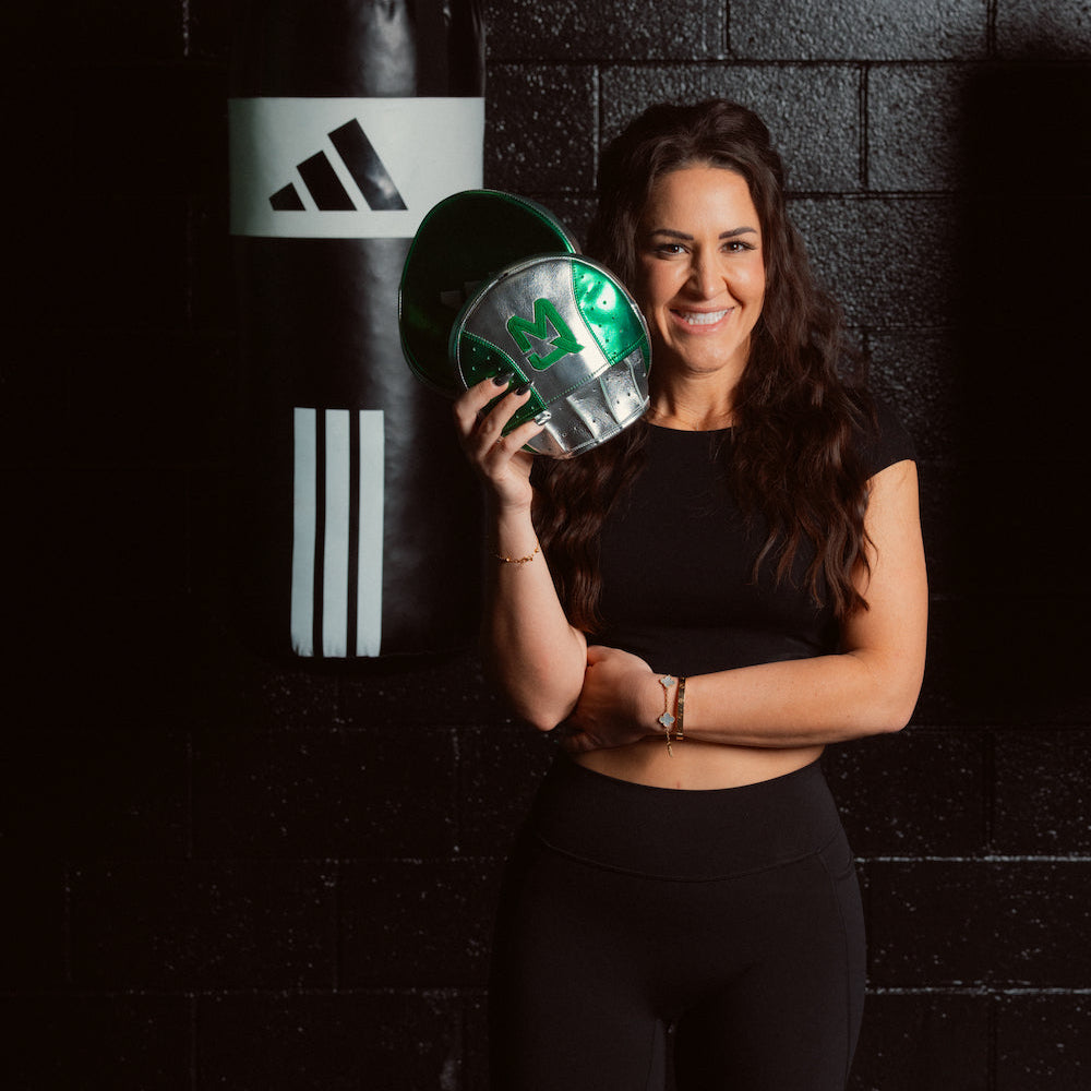A woman in a black athletic outfit stands in front of a black brick wall, holding a green and silver boxing glove. A black Adidas punching bag hangs on the wall behind her.